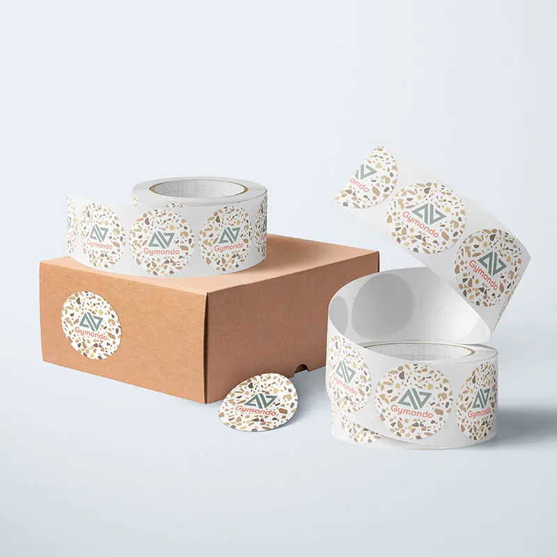 A brown cardboard box is sitting on a light grey surface. It has circular stickers with a decorative pattern of multicolored leaves and the text "Gumtoo." Next to the box, there are two rolls of similar stickers, with one roll unwrapping slightly in this cozy home setting.