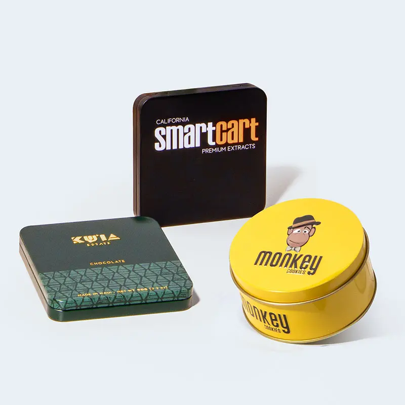 Three distinct product tins are displayed: a square, brown "Smartcart" tin for premium extracts, a green and gold "Kiva" tin for chocolates, and a round, yellow "Monkey" tin with a cartoon monkey design. These charming tins could easily find a place in any home against the plain, light background.